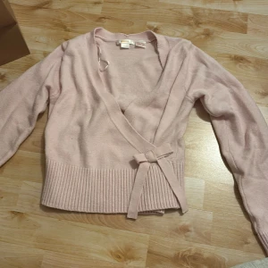 Ljusrosa omlottkofta från Gina Tricot - Supersöt ljusrosa omlottkofta från Gina Tricot med knytband i midjan och bred ribbad mudd nedtill. Koftan har lång ärm och är perfekt för lager-på-lager-stil. Mjuk och skön, passar perfekt till både jeans och kjol.