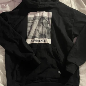 Svart hoodie från Saligia med tryck - Svart hoodie från Saligia med stor vit text 'Rich in Experience' på ryggen och grafiskt tryck framtill. Klassisk känguruficka, huva och långa ärmar. Mjuk och skön i bomullsmix, perfekt för chill dagar.