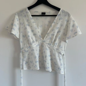 Blommig omlottopp från Gina Tricot - Supersöt vit omlottopp från Gina Tricot med ljusblått blommönster. Toppen har v-ringning, korta ärmar och knytband i midjan som knyts som en rosett där bak. Väldigt bra skick!
