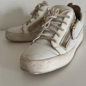 Vita sneakers från Giuseppe Zanotti - Säljer ett par vita sneakers från Giuseppe Zanotti med guldfärgade dragkedjor och logga. Skorna är i skinn och har snörning samt rund tå. Snygg och chunky sula som ger en cool vibe. Perfekta för dig som gillar exklusiva detaljer och italiensk design. Skorna följer inte med og det är därför den säljs billigt… och skorna blir rengjorda innan dom skickas ut 