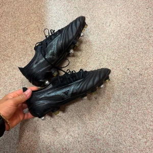Mizuno fotbollsskor SG  - Säljer ett par svarta Mizuno fotbollsskor med snörning och metall-dobbar. Skorna har en stilren design med diskreta detaljer och är tillverkade i skinn och syntet. Perfekta för matcher på gräsplan. Snygg och klassisk look för dig som gillar kvalitet. Storlek 41
