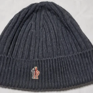 Mörkgrå ribbad mössa från Moncler - Snygg mörkgrå ribbstickad mössa från Moncler med klassisk logotyp framtill. Mössan är tillverkad i mjuk ull och har en enkel, tidlös design som passar perfekt för kyliga dagar. Perfekt accessoar för dig som gillar stilrena och exklusiva märken. Äkta. 