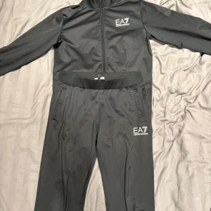 Svart EA7 tracksuit Emporio Armani - Säljer en svart tracksuit från EA7 Emporio Armani, storlek 14A (164 cm). Setet består av zip-jacka med ståkrage och matchande byxor, båda med EA7-logga i vitt. Materialet är mjukt och stretchigt, perfekt för chill eller sport. Snygg och stilren design med bekväm passform.