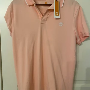 Ljusrosa Timberland piké slim fit - Fräsch ljusrosa pikétröja från Timberland i slim fit-modell. T-shirten har korta ärmar, vit broderad logga på bröstet och vit kant på kragen. Tillverkad av 100% ekologisk bomull, perfekt för dig som gillar stilren och enkel design.