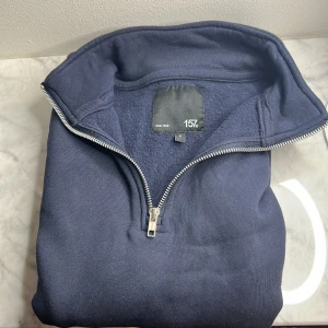 Marinblå half zip tröja från 157 - Mysig marinblå långärmad tröja från 157 med half zip-dragkedja och hög krage. Insidan är fleecefodrad för extra värme och komfort. Perfekt för kyliga dagar och enkel att matcha med jeans eller mjukisbyxor.