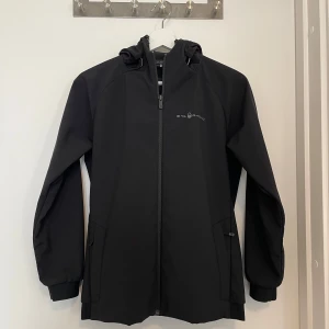 Svart vindjacka från Sail Racing - Svart vindjacka från Sail Racing med huva och dragkedja framtill. Jackan har diskreta loggor på bröstet, ärmen och huvan. Tillverkad i ett slätt syntetmaterial och har två fickor med dragkedja. Perfekt för blåsiga dagar och snygg, clean look.