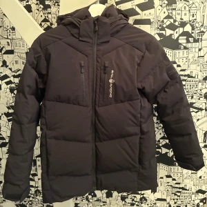 Svart Sail Racing dunjacka med huva - Svart dunjacka från Sail Racing, modell Antarctica Expedition. Jackan har huva, två bröstfickor med dragkedja och Sail Racing-logga på bröstet. Tjock och varm med quiltad design, perfekt för kalla dagar. Badge på ärmen och snygga detaljer, och även en bra innerficka. Jackan är i barnmodell och passar någon som är omkring 150cm😁