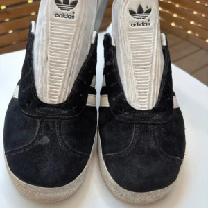 Adidas Gazelle svartvita sneakers - Klassiska Adidas Gazelle sneakers i svart mocka med vita detaljer och de ikoniska tre ränderna på sidan. Skorna har rund tå, platt sula och vit logga på tungan och hälen. Perfekta för en stilren och sportig look.