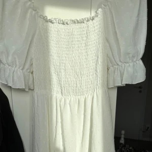 Vit medellång klänning med puffärm och knyt - Superfin vit klänning med puffärmar och smockad överdel. Klänningen har volangdetaljer vid ärmslut och en snygg knytning i ryggen som ger en öppen rygg. Tunt, lätt tyg med diskret struktur och romantisk känsla. Aldrig haft chansen o använda den