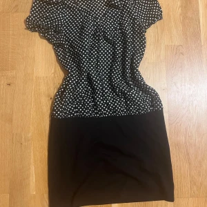 Svart prickig kortklänning La Chapelle - Kort svart klänning från La Chapelle med vitprickigt mönster upptill och enfärgad svart kjol. Klänningen har v-ringning, korta ärmar och markerad midja. Perfekt för dig som gillar retrovibbar och klassisk stil. Passar XS och S