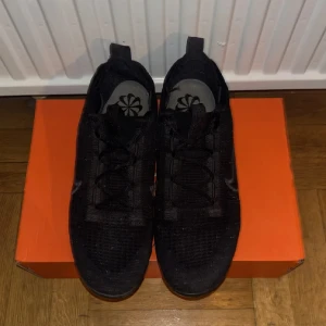 Svarta Nike Vapormax sneakers - Säljer ett par helsvarta Nike Vapormax sneakers med stickad ovandel och klassisk Swoosh på sidan. Skorna har snörning och en tydlig platt sula med den ikoniska Vapormax-luftbubblan. Perfekta för dig som gillar sportig och stilren look.