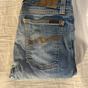 Blåa nudie jeans - Snygga ljusblå jeans från Nudie Jeans med klassisk femficksdesign och gul kontrastsöm på bakfickan. Jeansen har smal passform och är tillverkade i mjukt denimtyg med dragkedja och knapp framtill. Perfekta för en avslappnad och trendig look.