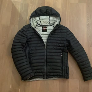 Svart dunjacka med huva från Colmar - Mörkblå dunjacka från Colmar Originals med horisontella sömmar, huva och dragkedja framtill. Jackan har två sidofickor med dragkedja och en ljusgrå insida. Jackan har minimala repor på höger arm(bild 4) men är inte något som märks. Perfekt nu för hösten. Skriv vid intresse!
