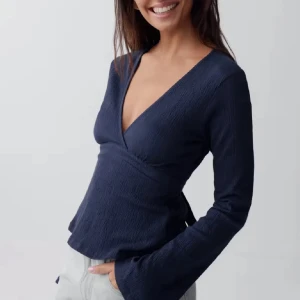 Mörkblå omlottopp med knyt i midjan - Snygg mörkblå omlottopp från Gina Tricot med v-ringning och långa ärmar. Toppen har knytband i midjan som ger en fin siluett och är tillverkad i ett mjukt, lätt strukturerat material. Perfekt för dig som gillar stilrena och trendiga plagg.