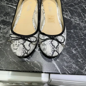 Ballerinaskor i ormmönster från KappAhl - Snygga ballerinaskor från KappAhl i svartvitt ormmönster med rund tå och dekorativ rosett framtill. Skorna har svarta kanter och platt sula.