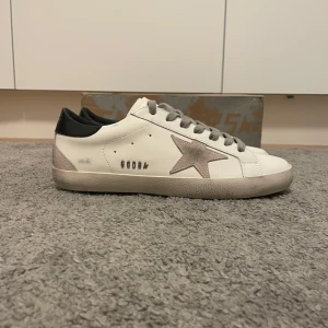 Golden Goose Superstar sneakers vit/grå - Golden Goose Superstar sneakers i vitt skinn med grå mockadetaljer och grå snörning. Skorna har en ikonisk stjärna i mocka på sidan, svart hälparti och platt sula. Snygg och avslappnad look med unika detaljer, perfekt för dig som gillar streetstyle.