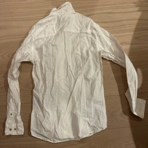 Vit långärmad skjorta i bomull - Klassisk vit skjorta med lång ärm och knappar framtill. Skjortan har enkel krage och är tillverkad i mjuk bomull, perfekt för en clean och stilren look. Passar dig som gillar basic och tidlös stil.