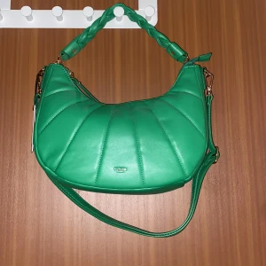 Grön axelväska från Ulrika Sweden - Snygg grön axelväska från Ulrika Sweden med quiltad framsida och flätat handtag. Väskan har guldfärgade detaljer, justerbar axelrem och dragkedja både upptill och på baksidan. Tillverkad i syntetmaterial och har en modern, rundad form.