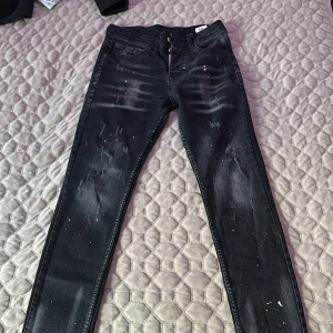 Svarta skinny jeans från Dsquared2 - Säljer ett par svarta skinny jeans från Dsquared2 med coola slitningar och färgstänk i vitt och rött. Jeansen har knappgylf, klassisk femficksdesign och är tillverkade i bomull och polyester. Perfekta för dig som gillar en edgy streetstil.