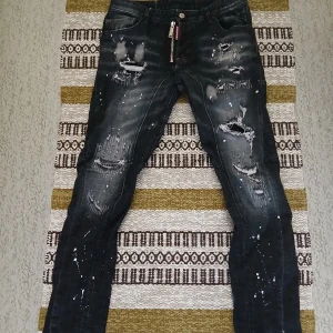 Svarta Dsquared2 jeans med slitningar - Snygga svarta jeans från Dsquared2 med coola slitningar och färgstänk. Modellen har smal passform och klassisk femficksdesign. Jeansen har dragkedja och tydlig Dsquared2-logga vid gylfen. Perfekta för dig som gillar en edgy streetstil. Storlek 44 men passar 40.
