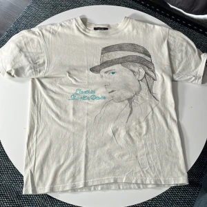 Vit t-shirt med 80-talsprint Global Work - Säljer en vit t-shirt från Global Work i bomull med coolt 80-talsinspirerat tryck och tecknad profil på framsidan. Turkos textdetalj på både fram- och baksida. Klassisk passform och korta ärmar, perfekt för dig som gillar retro vibes.