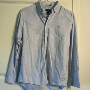 Ljusblå skjorta från GANT, Oxford - Klassisk ljusblå skjorta från GANT, modell The Archive Oxford. Skjortan har button-down krage, broderad logga på bröstet och knäppning framtill. Tillverkad i bomull och har långa ärmar. Perfekt för en clean och stilren look. xs-164 ~ 13-14y