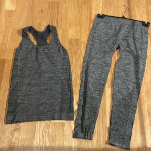 Grå träningstopp i syntetmaterial - Säljer en grå, ärmlös träningstopp med brottarrygg, märke Panos Emperio. Toppen är tillverkad i ett stretchigt syntetmaterial som andas och har en sportig look med diskret mönster längs sidorna. Perfekt för gymmet eller löprundan.