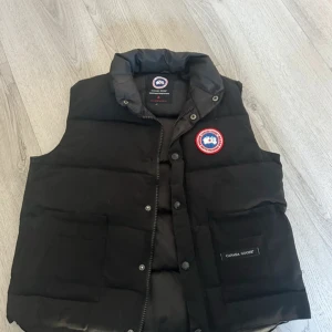 Svart dunväst från Canada Goose s/xs - Svart dunväst från Canada Goose med klassisk logga på bröstet. Västen har hög krage, två stora fickor framtill och stängs med både dragkedja och knappar. Perfekt för lager-på-lager och riktigt snygg till streetwear.
