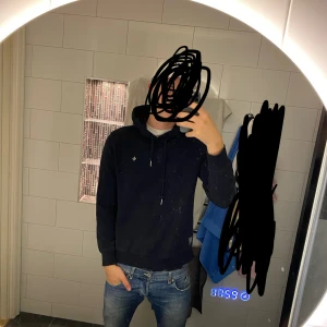 Morris hoodie  - Marin blå hoodie från Morris med klassisk huva och snörning. Tröjan har en liten broderad logga på bröstet och en patch nertill. Tillverkad i mjuk bomull, perfekt för chill dagar. Snygg och enkel att matcha med jeans eller joggers. Hör av dig vid fler funderingar!