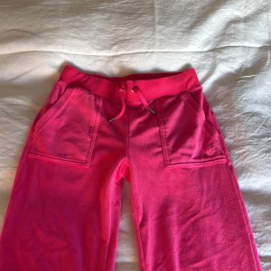Juicy Couture rosa mjukisbyxor XS - Juicy Couture mjukisbyxor i stark rosa färg med bred resår och snörning i midjan. Byxorna har raka ben, stora fickor fram och bak samt broderad JC-logga på ena fickan. Mjukt material som känns skönt mot huden.