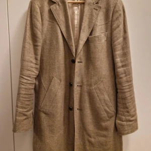 Beige kappa från The Tailoring Club - Snygg beige kappa från The Tailoring Club i en mix av 75% hampa och 25% ull. Klassisk design med tre knappar, breda slag och stora fickor framtill. Fodrad med viskos för extra komfort. Perfekt för dig som gillar stilrena och tidlösa plagg.