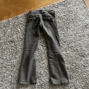 Grå bootcut jeans från Gina Tricot - Snygga grå jeans från Gina Tricot i storlek 146. Modellen har bootcut-ben och klassiska fem fickor, inklusive ficklock bak. Jeansen har en cool tvättad look och är tillverkade i mjukt denimtyg med dragkedja och knapp framtill.
