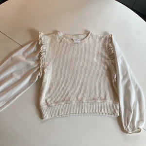 Beige ribbad tröja med volang Zara - Supersöt beige ribbad tröja från Zara med långärmade släta ärmar och volangdetaljer vid axlarna. Tröjan har rund halsringning och är i mjuk bomullsmix. Perfekt för dig som gillar en stilren men ändå söt look.