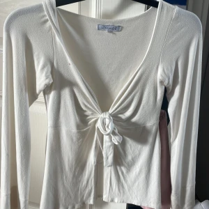 Vit omlottopp med knyt från Design by Si - Supersöt vit omlottopp från Design by Si i storlek XS/S. Toppen har långa ärmar, djup v-ringning och snygg knytning framtill. Perfekt för dig som gillar en feminin och trendig look. Mjuk och skön i bomullsmix. Liten fläck vid typ halsen men ingenting man ser när man väl har på sig den❤️