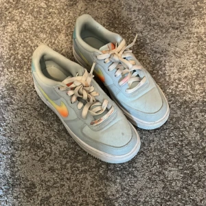 Nike Air Force 1 med färgad swoosh - Nike Air Force 1 sneakers i ljusblått med en snygg swoosh i gradient från gult till orange. Klassisk siluett med snörning och platt sula. Ovandel i syntet och skinn, perfekt för dig som gillar färgdetaljer på dina sneakers.