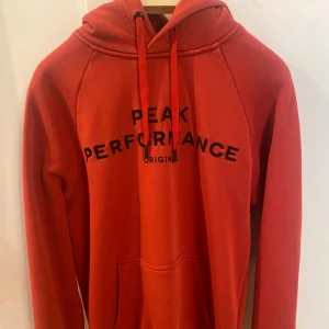 Röd hoodie Peak Performance M - Snygg röd hoodie från Peak Performance i storlek M. Tröjan har svart tryck på bröstet, huva med snörning och en stor magficka. Perfekt för chill dagar och har en klassisk, sportig look. Materialet är mjuk bomull som känns skönt mot huden.