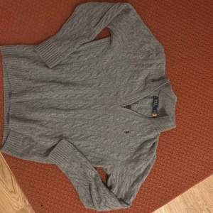 Grå kabelstickad halfsip tröja Polo Ralph Lauren - Kabelstickad grå tröja från Polo Ralph Lauren med v-ringning och broderad logga på bröstet. Tröjan har långa ärmar och ribbade muddar vid ärmslut och nederkant. Perfekt för lager-på-lager och klassisk stil.
