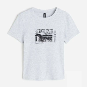 Ljusgrå t-shirt med New York tryck - Säljer en ljusgrå t-shirt från Divided med ett coolt svart tryck av en pizzeria i Downtown, New York på bröstet. T-shirten har rund hals och korta ärmar, perfekt för dig som gillar streetstyle och grafiska prints. Det står M men passar som storlek S eftersom den är väldigt tajt!