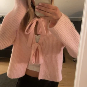 Rosa stickad kofta med rosetter - Supergullig ljusrosa stickad kofta med två stora rosetter framtill. Koftan har lång ärm och är lite croppad i modellen. Perfekt för dig som gillar en söt och trendig look. Mjuk och mysig att ha på sig.