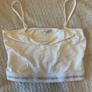 Vit linnetopp från Calvin Klein Jeans - Säljer en vit linnetopp från Calvin Klein Jeans med tunna axelband och logotyp längs nederkanten. Toppen har en enkel och stilren design, perfekt att matcha med jeans eller kjol. Mjuk bomullskänsla och croppad passform.