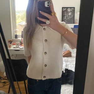 Vit stickad kofta från Gina Tricot - Superfin vit stickad kofta från Gina Tricot med kort ärm och stora knappar framtill. Koftan har en klassisk krage och ribbade kanter nedtill och vid ärmslut. Perfekt att slänga på sig över ett linne eller t-shirt för en chill look.