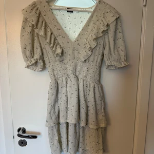 Blommig volangklänning Gina Tricot M - Supersöt kortklänning från Gina Tricot i ljusbeige med små gröna blommor. Klänningen har v-ringning, volangdetaljer längs urringningen och ärmarna, samt smock i midjan. Puffiga kortärmar och dubbla volanger i kjolen ger en romantisk vibe.