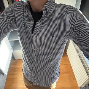 Ralph Lauren skjorta  - Säljer en slim fit skjorta från Ralph Lauren i vitt och blått med klassiska vertikala ränder. Skjortan har button down-krage, långa ärmar och broderad logga på bröstet. Tillverkad i bomull och stängs med knappar framtill.