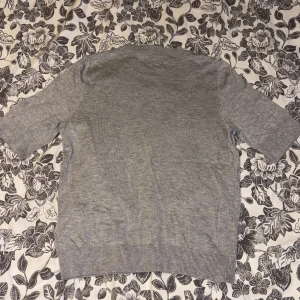 Grå stickad tröja med kort ärm - Säljer en stilren grå stickad tröja. Tröjan har rund halsringning, ribbade muddar och korta ärmar. Perfekt för lager-på-lager eller som den är under vår och sommar. 