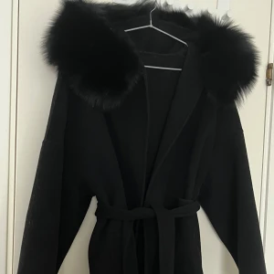 Svart kappa med på päls, bälte och fickor  - Svart kappa med stor huva kantad av fluffig päls. Jackan har bälte i midjan för snygg passform och långa ärmar. Perfekt för kalla dagar när du vill vara både varm och stilren. Den har även två fickor på framsidan. Använt ett par gånger men ser helt ny ut utan några skador. 
