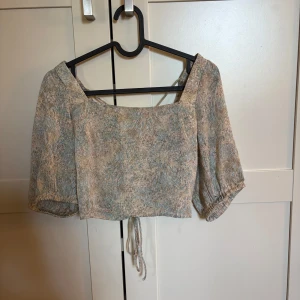 Beige och blå croppad blus med snörning - Säljer en croppad blus i beige och blått med abstrakt mönster. Toppen har puffiga korta ärmar, fyrkantig ringning och snörning i ryggen som ger en snygg detalj. Lätt och luftig, perfekt för varmare dagar.