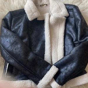 Svart teddyjacka från Altamira Milano - Snygg svart teddyjacka från Altamira Milano med vit fuskpäls på insidan, krage och ärmslut. Yttermaterialet har en cool skinnlook med struktur. Jackan har en klassisk, rak passform och är perfekt för kalla dagar.