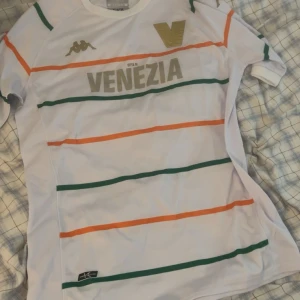Venezia FC vit fotbollströja Kappa M - Snygg vit fotbollströja från Venezia FC med gröna och orange horisontella ränder, guldiga detaljer och Kappa-logga på bröstet. Tillverkad i lätt och ventilerande material, perfekt för match eller träning. Klassisk passform och korta ärmar.