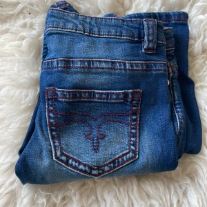 Blå jeans från Gina Tricot med röd söm - Snygga blå jeans från Gina Tricot i storlek 170. Jeansen har röda kontrastsömmar och cool broderad bakficka. Klassisk femficksmodell med dragkedja och knapp framtill. Perfekta för dig som gillar detaljer och en trendig look.