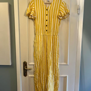 Gul- och vitrandig jumpsuit Lindex - Superfin jumpsuit från Lindex i gult och vitt med vertikala ränder. Klänningen har korta volangärmar, v-ringning och svarta knappar framtill. Tillverkad i ett lätt och skönt material, perfekt för varma dagar. Snygg och bekväm passform!